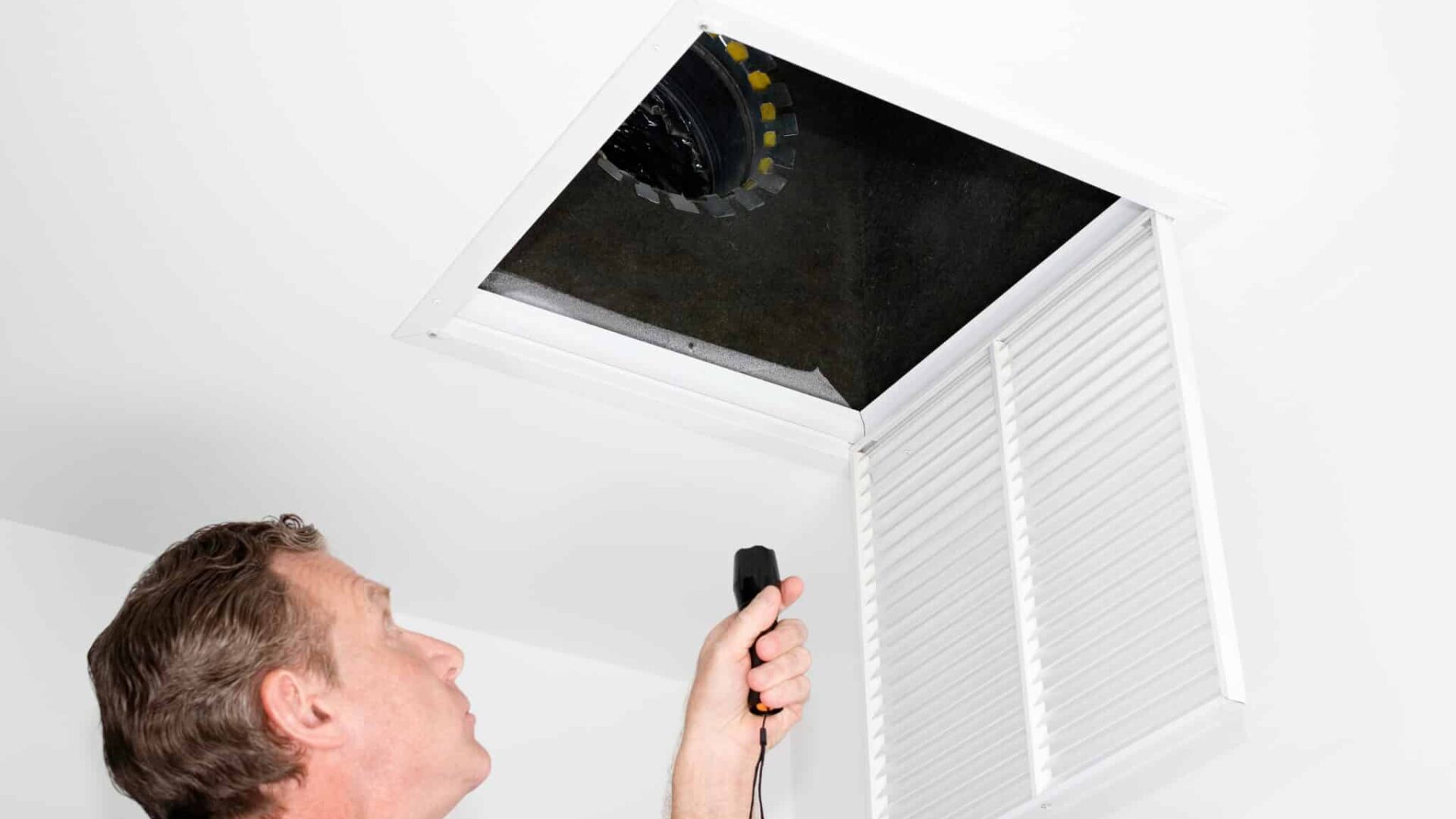 Male looking up into a ceiling air intake duct with a flashlight checking for maintenance. Person with a flashlight examining with a square opening of a home HVAC system
