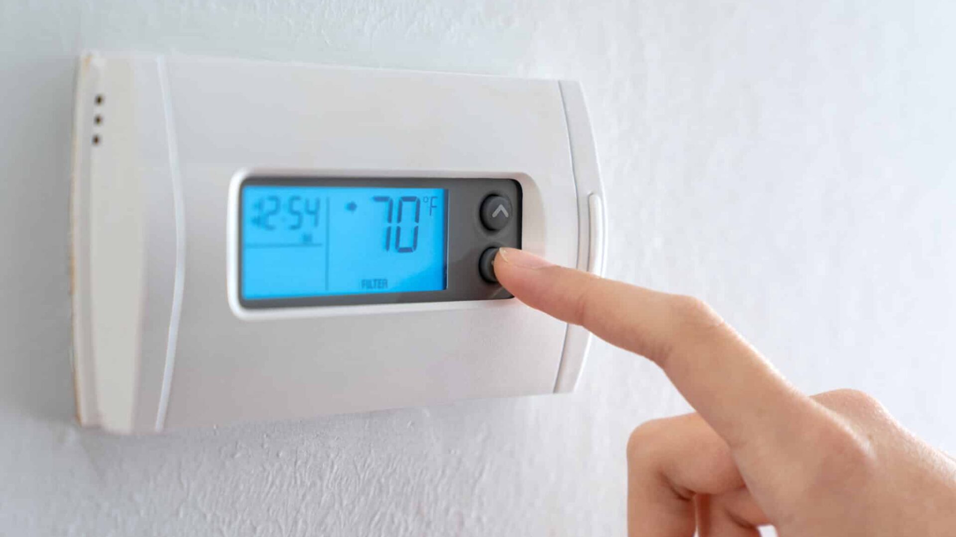 A woman is pressing the down button of a wall attached house thermostat with digital display showing temperature 70 degree Fahrenheit for heating, cooling, electricity and gas saving